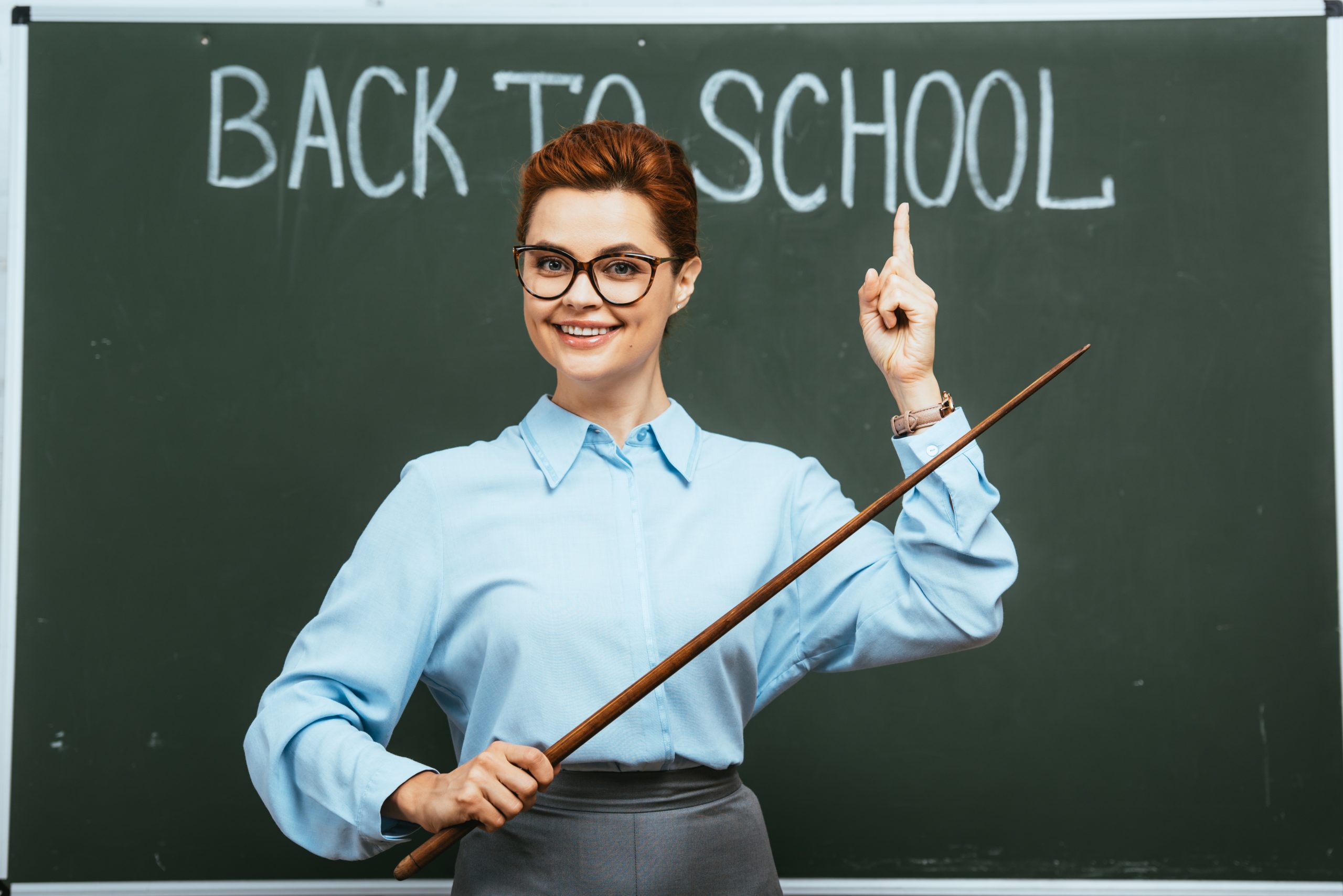 smiling teacher teaching Romanian A1 course and holding pointer and pointing with finger at back to school inscription on chalkboard