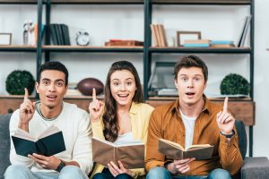 Three multiethnic students learn Romanian imperfect tense with books and pointing with fingers up in living room.
