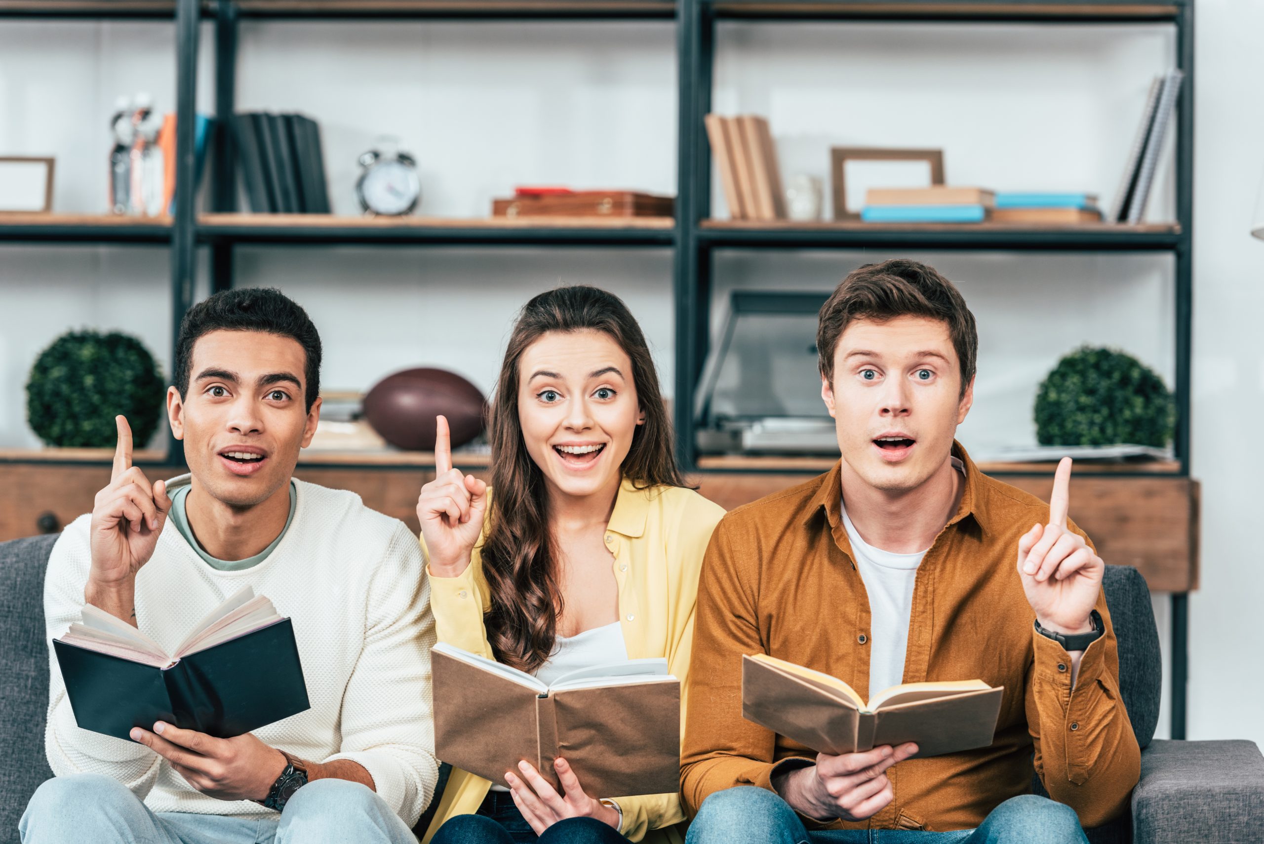 Three multiethnic students learn Romanian imperfect tense with books and pointing with fingers up in living room.